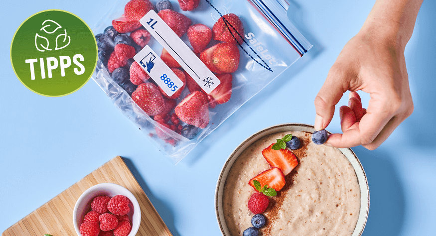 Hand garniert eine Schale Porridge mit frischen Beeren. Im Hintergrund liegt ein Gefrierbeutel mit gefrorenen Beeren auf einem hellblauen Tisch. Links ist ein Holzbrett mit einer kleinen Schale Himbeeren zu sehen. Hand garniert eine Schale Porridge mit frischen Beeren. Im Hintergrund liegt ein Gefrierbeutel mit gefrorenen Beeren auf einem hellblauen Tisch. Links ist ein Holzbrett mit einer kleinen Schale Himbeeren zu sehen.