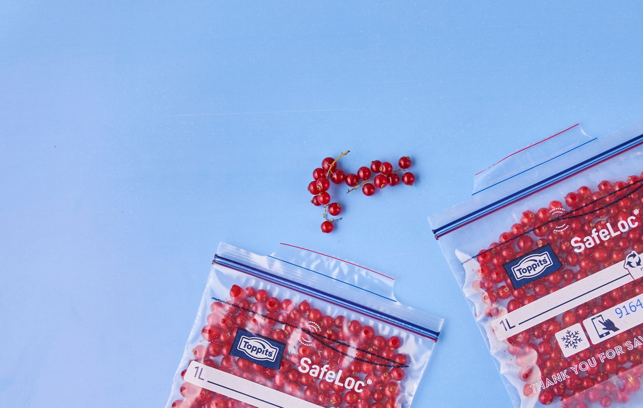 Zwei Toppits SafeLoc Gefrierbeutel mit eingefrorenen roten Johannisbeeren liegen auf einer blauen Fläche, daneben einige frische Johannisbeeren Zwei Toppits SafeLoc Gefrierbeutel mit eingefrorenen roten Johannisbeeren liegen auf einer blauen Fläche, daneben einige frische Johannisbeeren